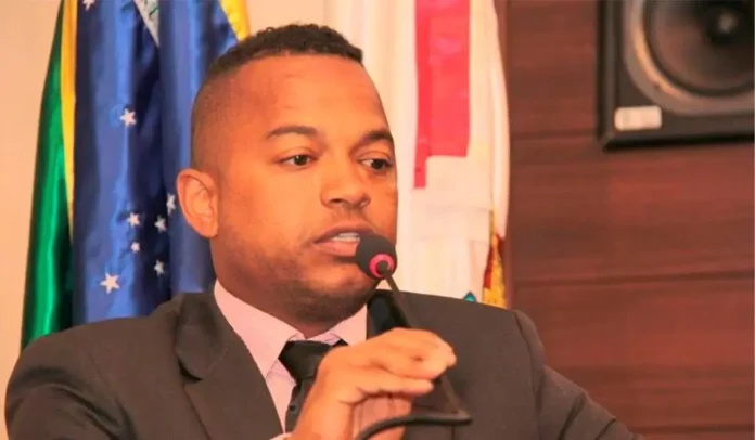 Pessoa falando ao microfone, com bandeiras ao fundo e painel de áudio na lateral.