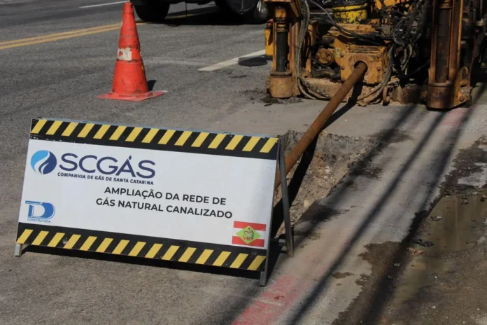 Placa de obras indicando a ampliação da rede de gás natural canalizado pela SCGÁS. Equipamento em atividade ao fundo.