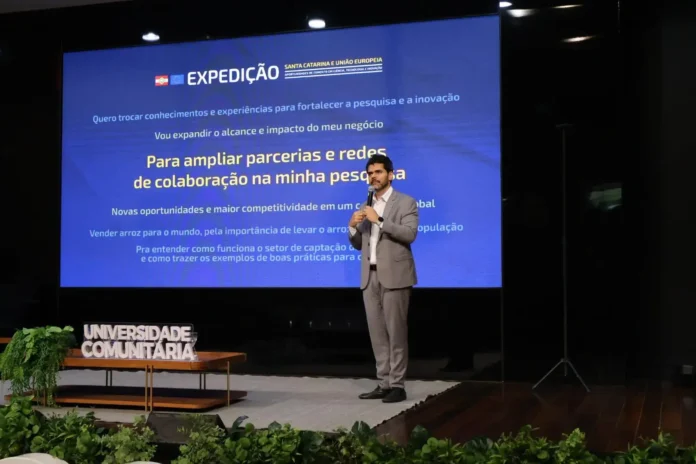 Homem em pé no palco durante evento, apresentando em frente a um telão com informações sobre pesquisa e colaboração. Texto visível em destaque.