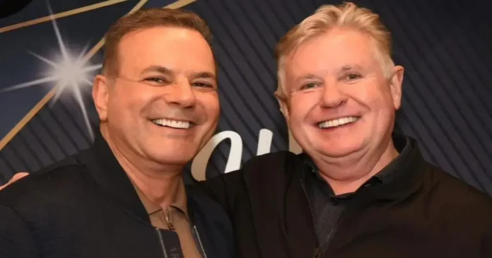 Dois homens sorrindo lado a lado, com fundo decorado e com brilho.