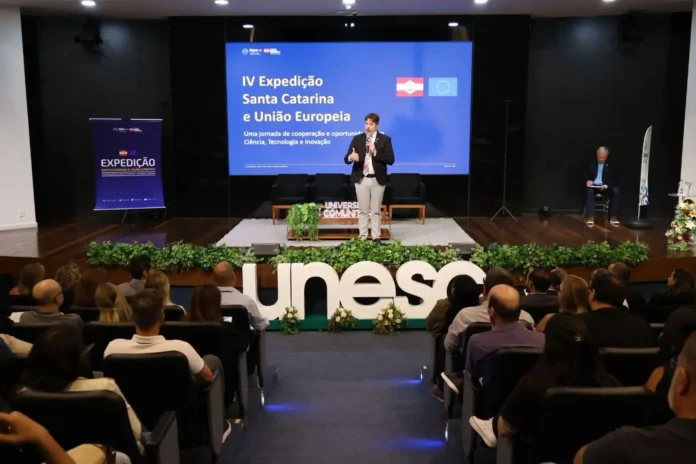 Evento com público assistindo a uma apresentação sobre a IV Expedição Santa Catarina e União Europeia, com destaque para o tema de ciência e inovação.