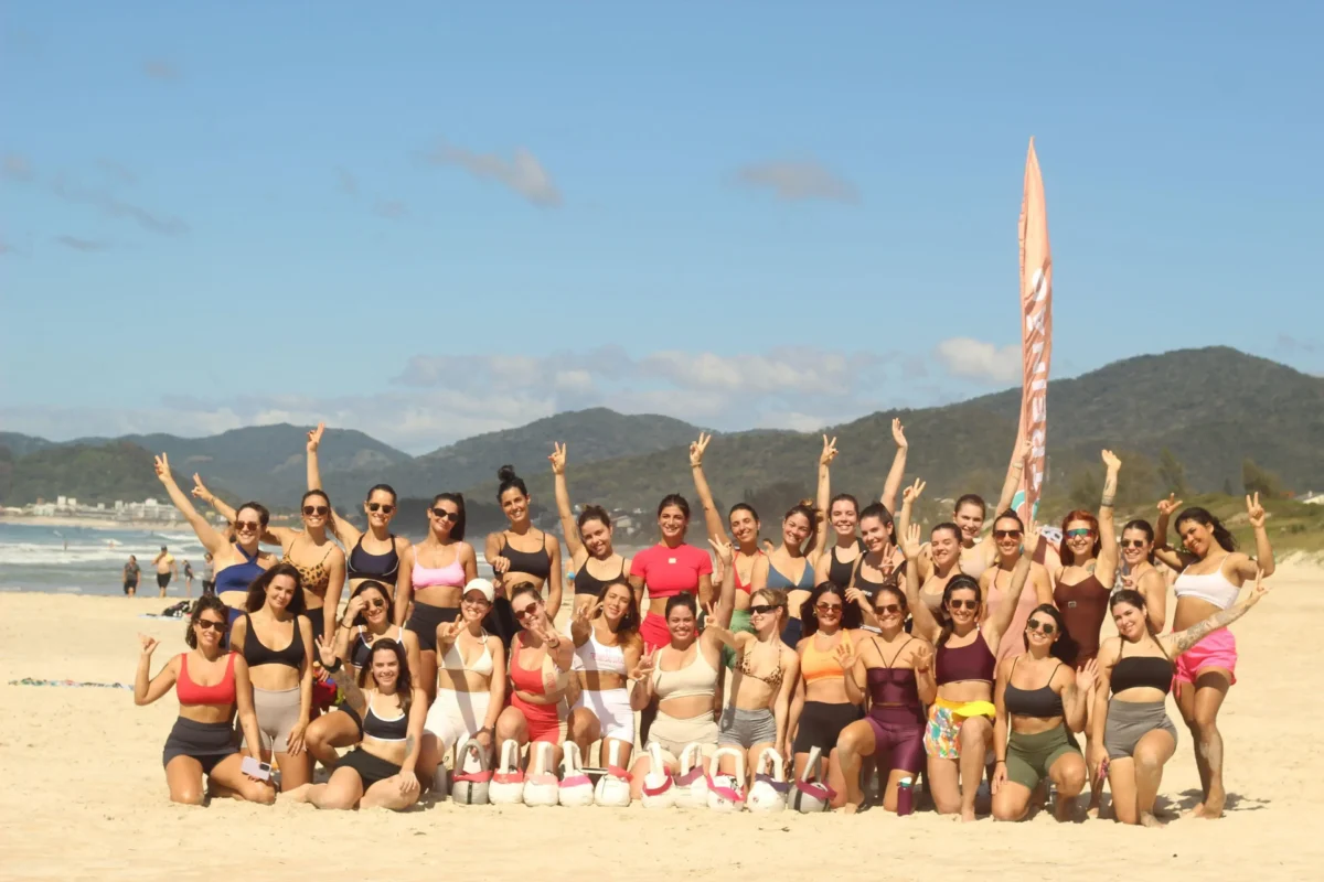 Grupo de mulheres em trajes de banho, com algumas fazendo sinal de paz, em uma praia com montanhas ao fundo.
