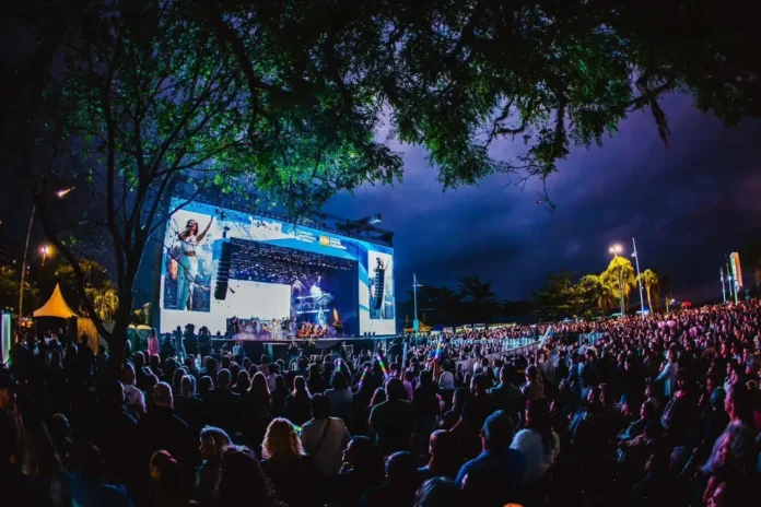 Grande multidão assistindo a um show ao ar livre, com palco iluminado e árvores ao redor.