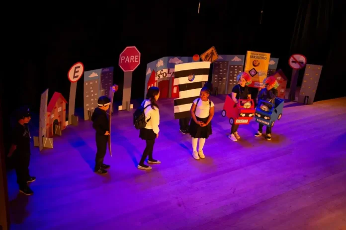 Crianças em apresentação teatral sobre segurança no trânsito, com cenário de cidade e placas de sinalização.
