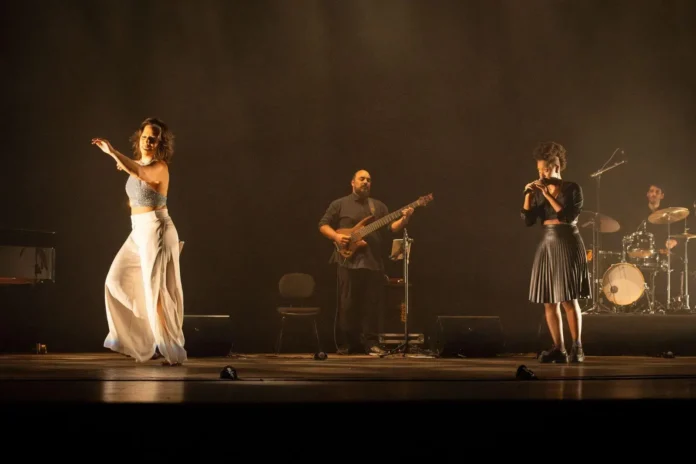 Performance musical no palco com quatro artistas, incluindo uma dançarina e músicos com instrumentos.