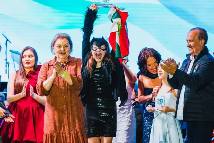 Jovem vestida de preto segura um troféu e uma bandeira, enquanto pessoas aplaudem ao fundo.