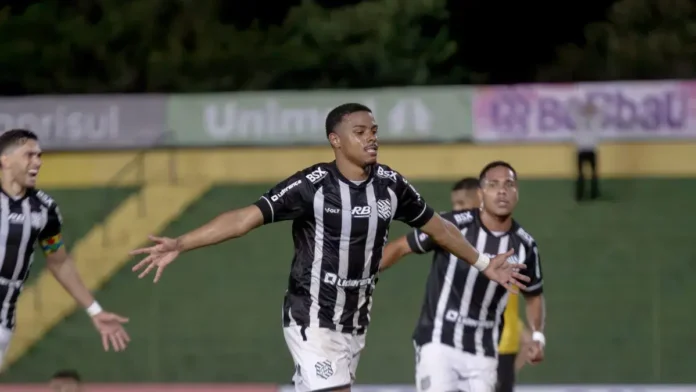 Jogador comemorando um gol em campo, cercado por colegas de time e com bandeira ao fundo.