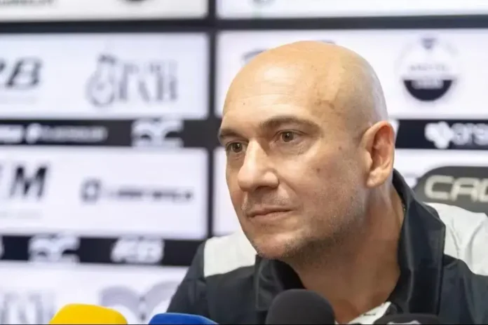 Homem calvo com expressão séria em um ambiente de coletiva de imprensa, com fundo de marcas e logotipos.