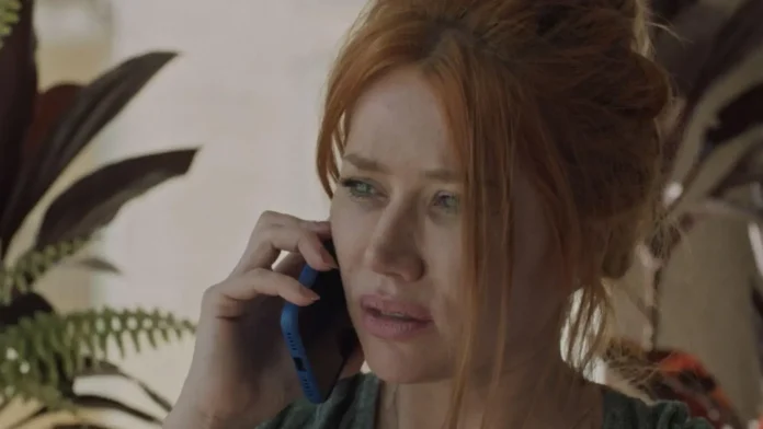 Mulher com cabelos ruivos falando ao telefone, visíveis folhas de plantas ao fundo.