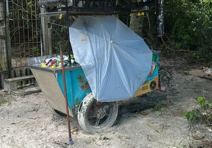 Carrinho de ambulante coberto com um guarda-sol, com latas visíveis, em área de areia e vegetação.