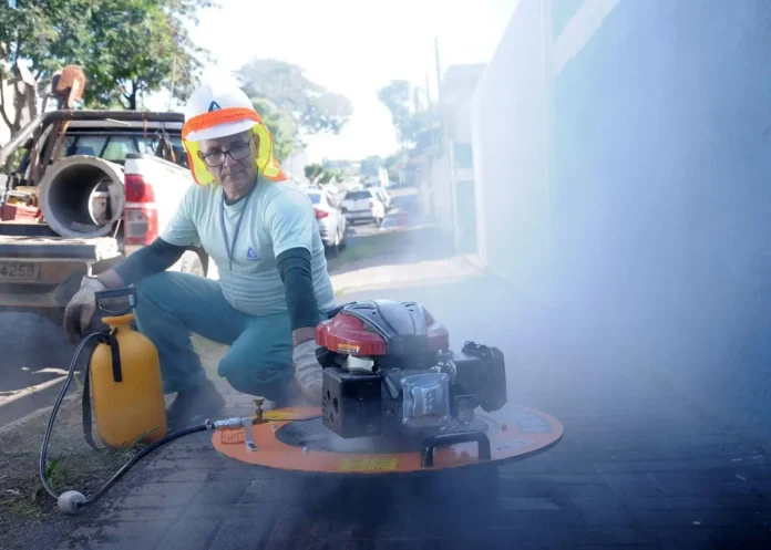 Homem utilizando um equipamento gerador de fumaça em uma calçada, com objetos e veículos ao fundo.