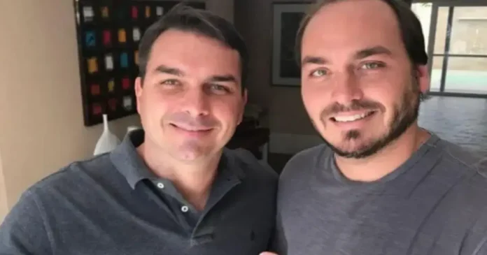 Duas pessoas sorrindo para a câmera em um ambiente interno, com uma parede decorada ao fundo.
