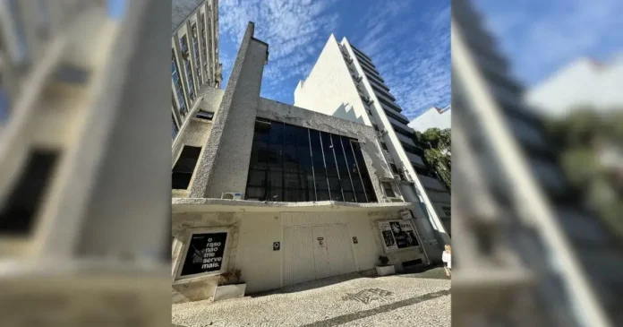 Fachada de um edifício com paredes de concreto, janelas grandes e cartazes na frente. Céu azul ao fundo.