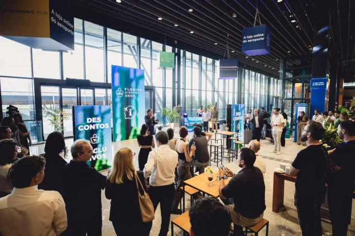 A área interna do aeroporto com pessoas assistindo a uma apresentação, painel digital visível no fundo.