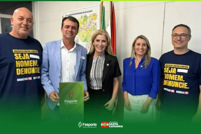 Grupo de pessoas em uma reunião com bandeiras ao fundo, segurando um cartaz da Fesporte. Texto visível: "SEJA HOMEM. DENUNCIE."