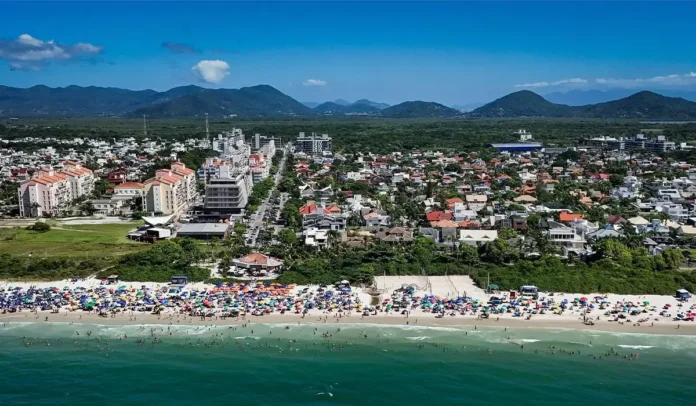 Praia com diversas pessoas, guarda-sóis e prédios ao fundo, cercada por montanhas sob céu azul.