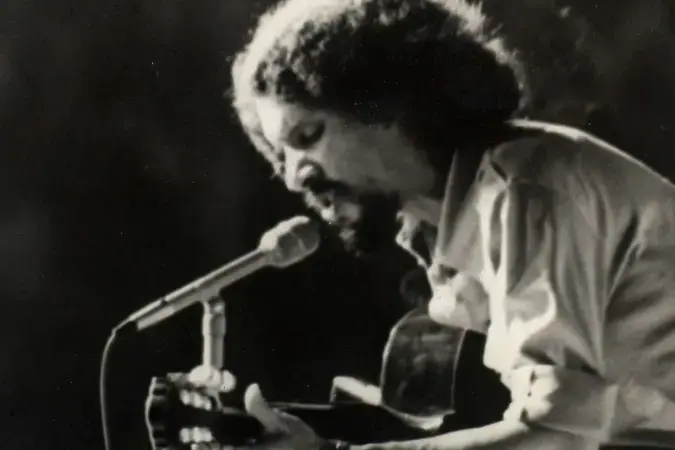 Músico tocando violão e cantando em frente a um microfone, retratado em preto e branco.