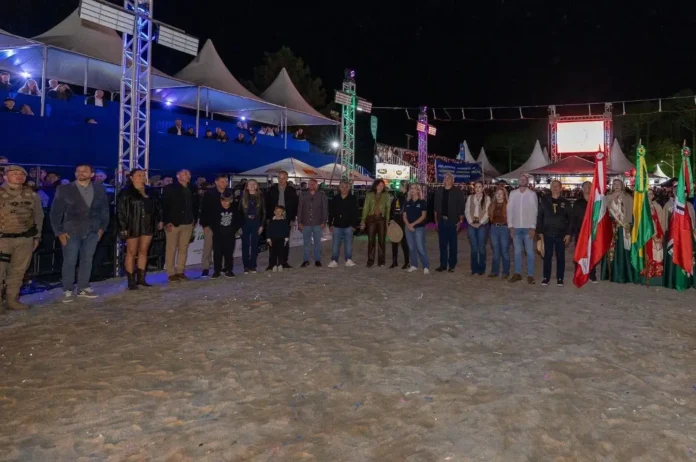 Grupo de pessoas em pé na areia, com bandeiras e barracas ao fundo sob luzes coloridas.