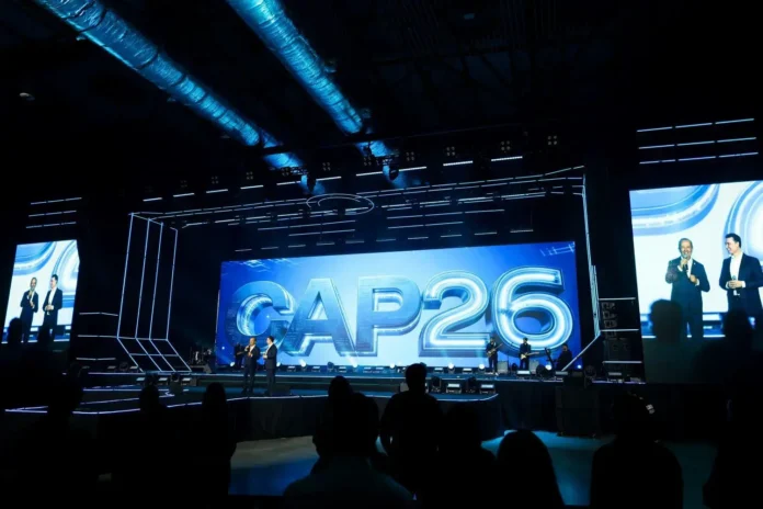 Dois apresentadores em um palco com um telão exibindo "CAP26". A iluminação do evento destaca a cena.