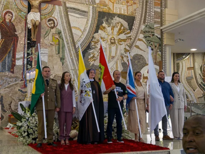 Grupo de pessoas em uma cerimônia, segurando bandeiras em frente a um fundo decorado com mosaicos religiosos.