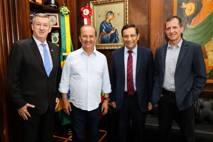 Quatro homens em um ambiente interno, com bandeiras ao fundo e uma imagem religiosa na parede.