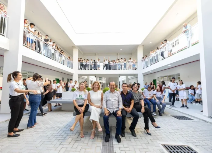 Grupo de pessoas sentadas em um banco no ambiente de uma escola, com alunos observando das varandas.