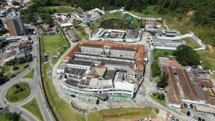 Vista aérea de um complexo prisional, com edifícios e áreas externas cercadas, próximo a uma estrada.