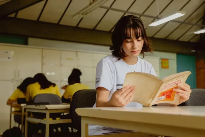 Estudante lendo um livro em uma sala de aula com mesas e cadeiras, ao fundo, outras pessoas com camisas amarelas.