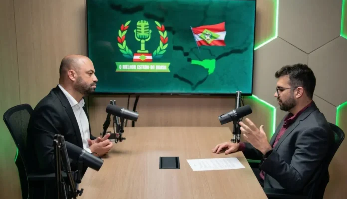 Dois homens sentados em uma mesa de gravação, discutindo, com microfones e uma tela ao fundo com o texto “O Melhor Estado do Brasil”.
