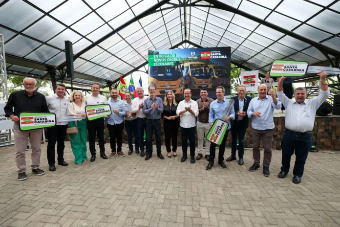 Grupo de pessoas em evento ao ar livre, segurando placas com o texto "Governo de Santa Catarina" e "Entrega de novos ônibus escolares". Banner ao fundo com ônibus.