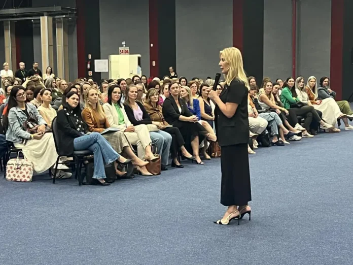 Cerca de cinquenta mulheres sentadas em cadeiras, assistindo a uma apresentadora que fala ao microfone em um evento.