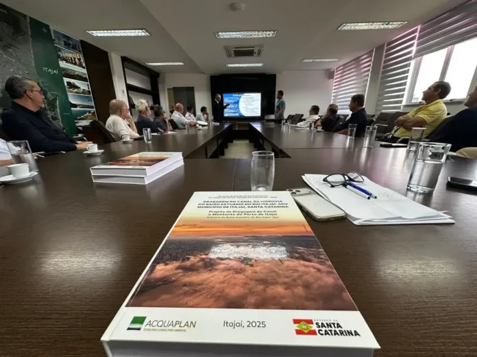 Reunião com diversas pessoas sentadas em torno de uma mesa, com um projeto na frente e uma apresentação ao fundo.