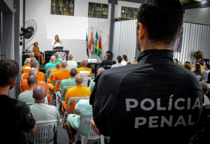 Reunião em penitenciária com palestras, assistentes e audiência de internos em cadeiras brancas.
