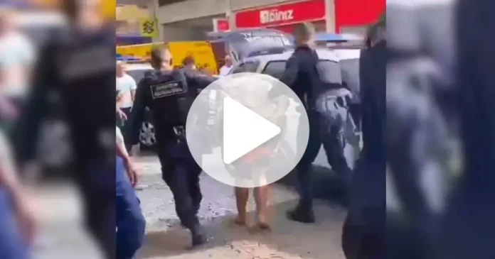 Grupo de pessoas em via pública observa abordagem policial a um homem, sob a presença de viaturas.