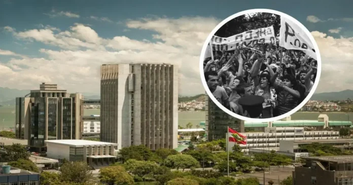 Vista de prédios e bandeira em meio a paisagem urbana, com grupo de pessoas segurando faixas em protesto.