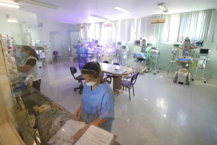 Ambiente hospitalar com profissionais de saúde, equipamentos médicos e uma mesa de trabalho.