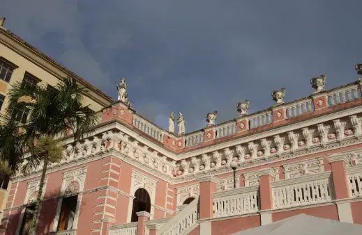 Construção histórica com detalhes em rosa, estátuas decorativas no telhado e vegetação ao redor.