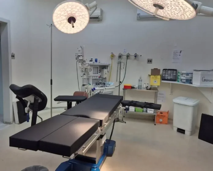 Sala cirúrgica com mesa de operação, iluminação cirúrgica, suporte para soro e equipamentos hospitalares ao fundo.