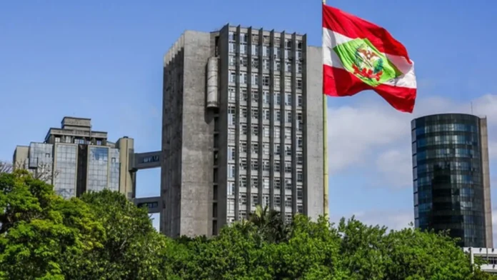 Bandeira do Peru hasteada em um mastro próximo a edifícios modernos e vegetação ao fundo.