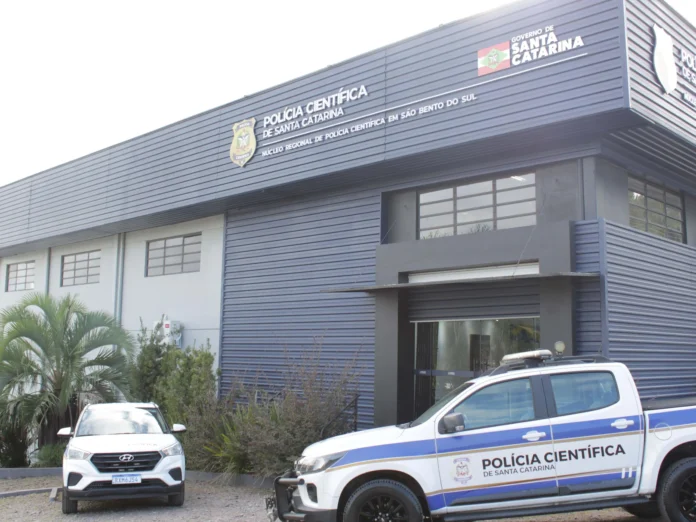 Prédio da Polícia Científica de Santa Catarina com veículos estacionados na frente e plantas ao redor.