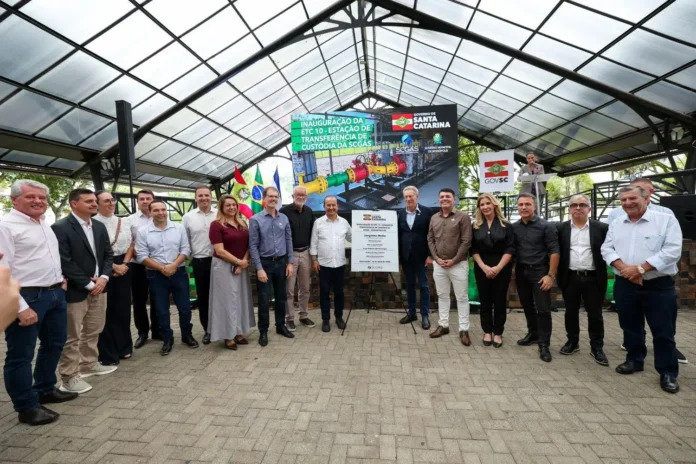 Grupo de pessoas em cerimônia de inauguração da Estação de Transferência de Custódia, com bandeiras ao fundo e painel informativo.