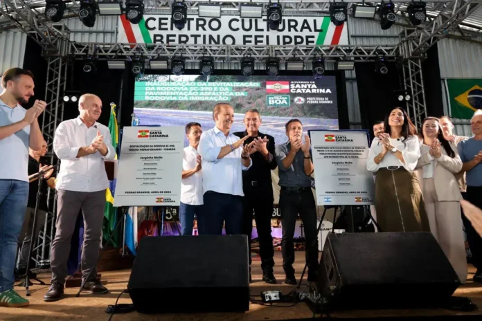 Cerimônia de inauguração com pessoas aplaudindo e placas sobre revitalização da rodovia SC-390, em um centro cultural.
