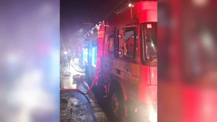 Caminhão de bombeiros com mangueira conectada, ao fundo, bombeiros e luzes de emergência visíveis.