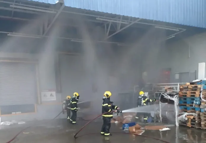 Bombeiros combatem incêndio em depósito, com equipamento de proteção e mangueiras, em meio a fumaça e paletes.