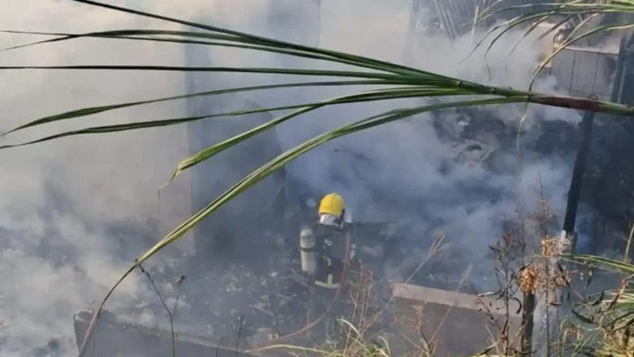 Bombeiro em ação combatendo incêndio em área de vegetação, cercado por fumaça e destroços.