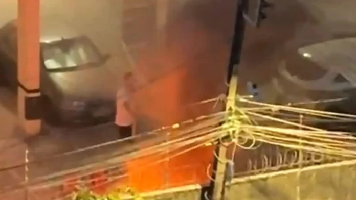 Homem próximo a veículo em chamas, enquanto fumaça densa se espalha pela área. Fios elétricos visíveis.