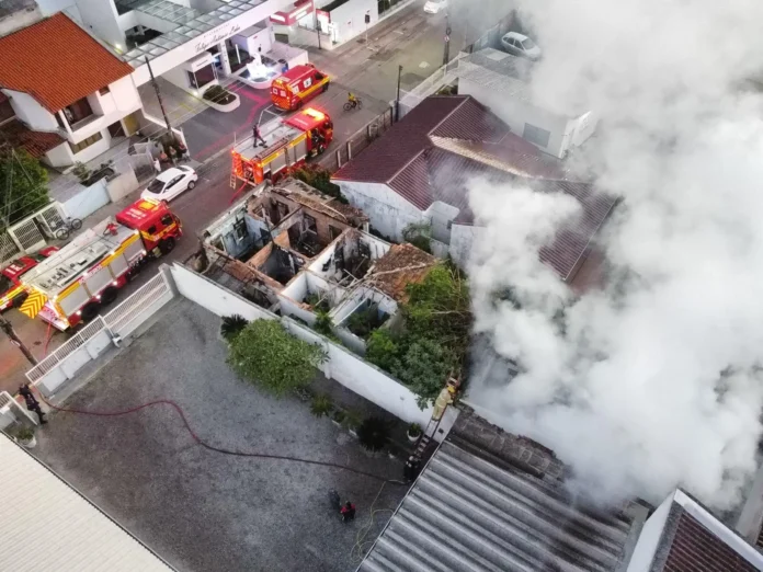 Incêndio em casa com fumaça saindo, viaturas do Corpo de Bombeiros e equipe no local realizando o combate ao fogo.