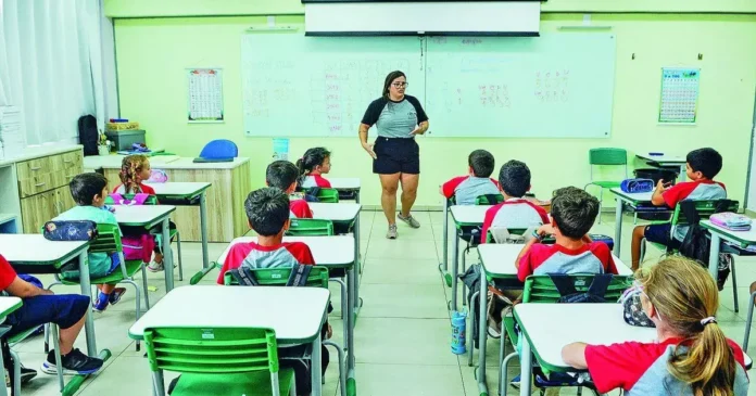 Sala de aula com alunos sentados em mesas, enquanto uma professora se dirige a eles.
