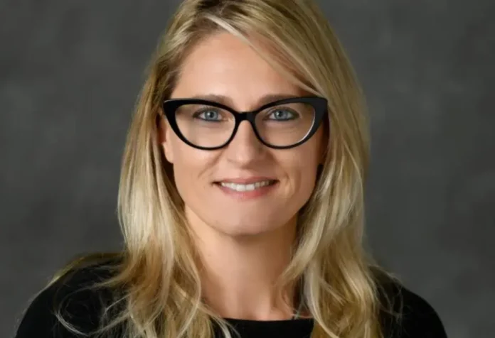 Retrato de uma mulher com cabelo loiro liso, usando óculos, sorrindo em fundo cinza.