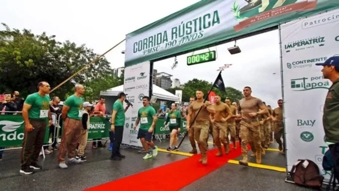 Participantes iniciam a Corrida Rústica em frente a um arco com o tempo marcado e público assistindo.